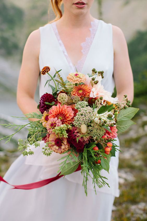 a creative fall wedding bouquet of orange and pink dahlias, burgundy ones, some seed pods and wildflowers, greenery and berries