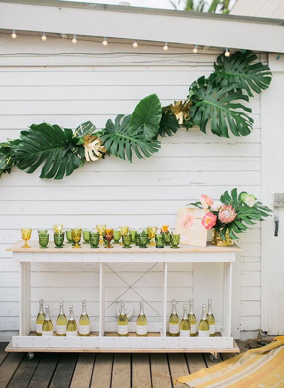 a fun tropical bridal shower drink station with colored glasses, lots of bottles and monstera leaf and king protea arrangements
