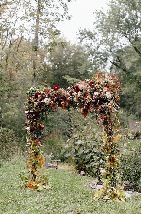 a gorgeous rustic fall wedding arch decorated with greenery and dried foliage, with bold and pastel dahlias and roses on top is very lush and eye-catchy