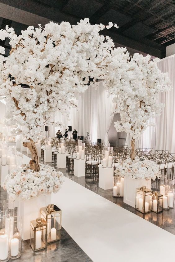 a jaw-dropping monochromatic wedding ceremony space with blooming cherry trees and neutral blooms, candle lanterns and white chairs