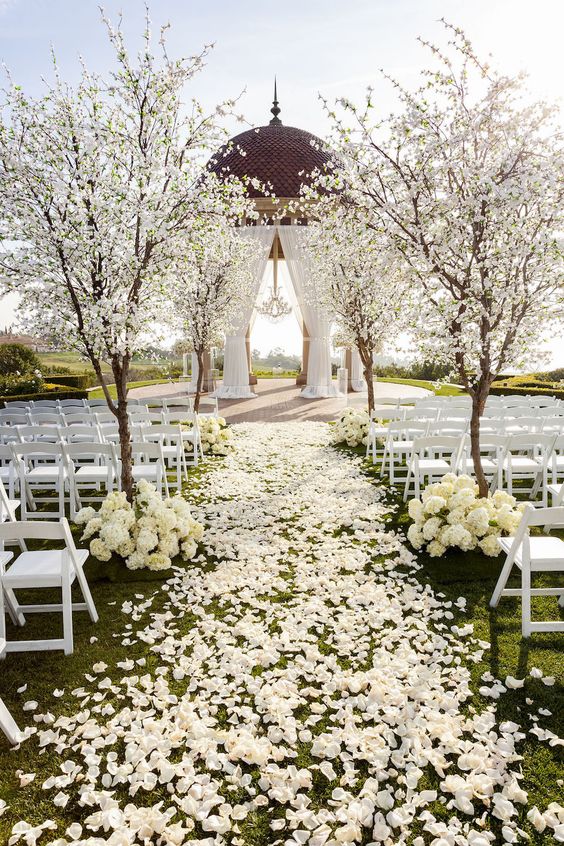 a jaw-dropping wedding ceremony space with white chairs, white petals on the ground, white cherry trees and an altar with curtains