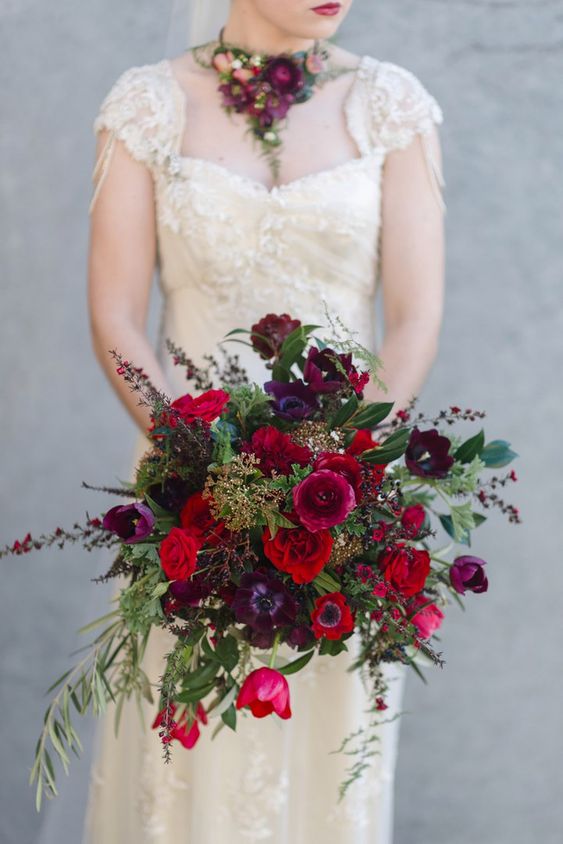 A jewel tone wedding bouquet of red anemones, burgundy and purple ranunculus and tulipes, greenery and bloomign branches