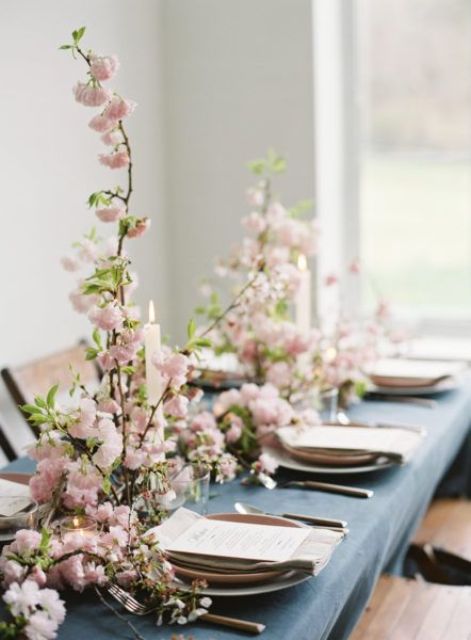 a lovely spring wedding tablescape with a blue tablecloth, pink cherry blossom and candles, neutral napkins and plates is beautiful