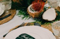 a tropical bridal shower tablescape with greenery, tropical blooms, coconuts, proteas, tropical leaves and wicker chargers