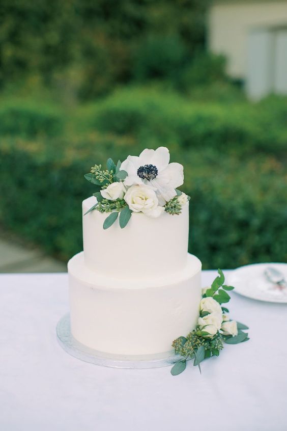 a white buttercream wedding cake topped with white blooms and an anemone looks ethereal and pretty