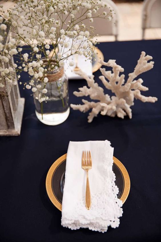 an elegant tablescape done with a navy tablecloth, corals, a gilded charger and cutlery, a baby's breath for a centerpiece