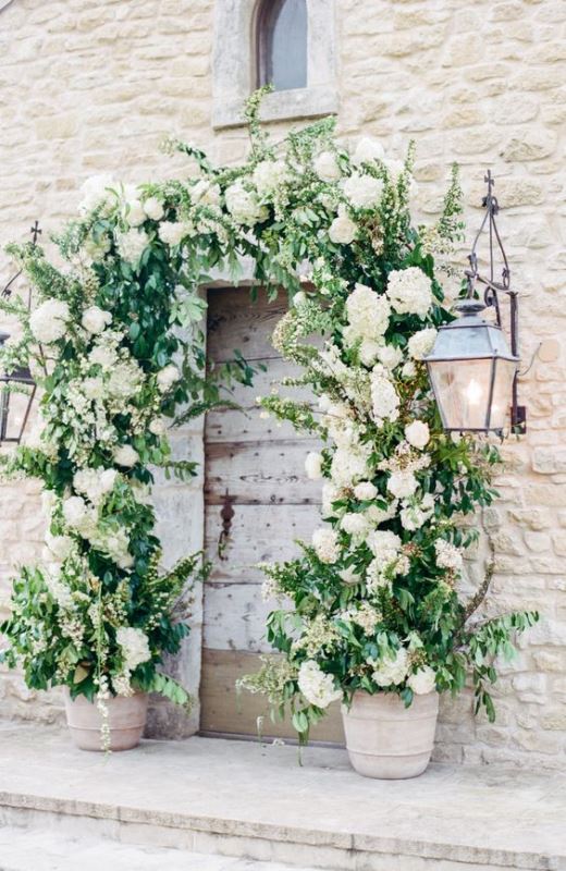 a wedding arch made of greenery and white blooms coming out of pots and climbing up the wall is a creative idea