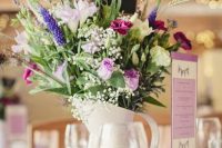 a bold wedding centerpiece of a neutral modern jug with bright and white blooms and lots of textural greenery plus a purple table name