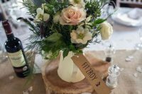 a pretty rustic wedding centerpiece of a neutral jug with pastel and neutral blooms and greenery, a wood slice and a cardboard tag is cute