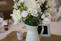a rustic vintage wedding centerpiece of a white metal jug with white blooms and greenery plus a burlap table runner is a cool idea
