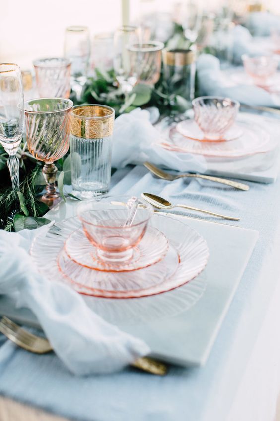 a beautiful mermaid bridal shower tablescape with a blue tablecloth and napkins, pink glass plates, a greenery runner and gold rim glasses