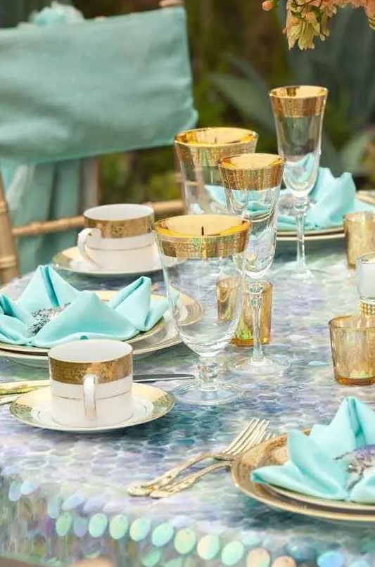 a bold mermaid bridal shower tablescape with a bright sequin tablecloth and turquoise napkins, gilded rim glasses and mugs