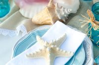 a bright mermaid bridal shower tablescape with a burlap runner, white and turquoise plates, seashells and starfish and blue jars