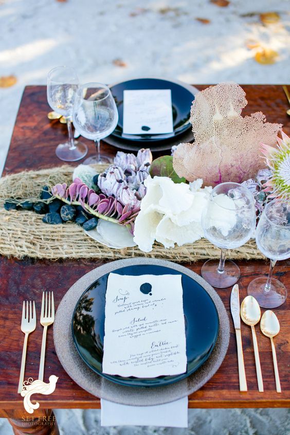 a bright mermaid-inspired tablescape with fishing net, purple and lilac blooms and pebbles, corals and beautiful dark green plates
