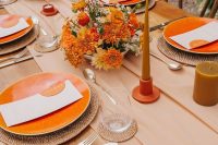 a bright retro bridal shower tablescape with a tan table runner, woven placemats, orange plates and rust candles, orange blooms and neutral cutlery