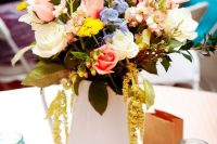a bright summer wedding centerpiece of a book stack, a white metal jug, bright blooms and greenery and candles in blue jars
