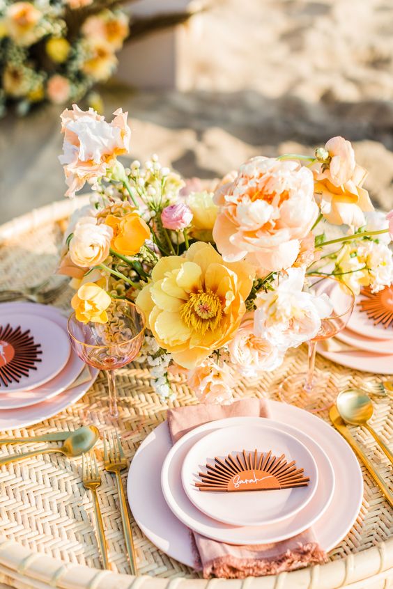a colorful retro bridal shower tablescape with a woven tablecloth, bold blooms, blush plates and gold cutlery and some pink glasses