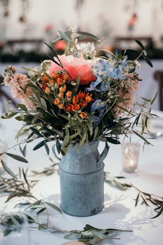 a colorful summer wedding centerpiece of a metal jug, pink, orange and blue blooms and lots of greenery for a rustic wedding