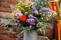 a colorful summer wedding centerpiece of a metal pitcher, bright blooms and greenery and much texture and dimension