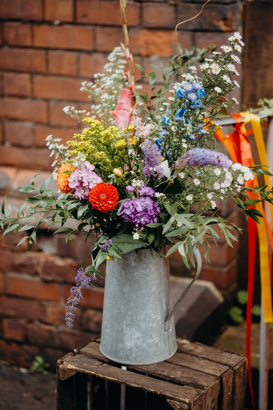 a colorful summer wedding centerpiece of a metal pitcher, bright blooms and greenery and much texture and dimension