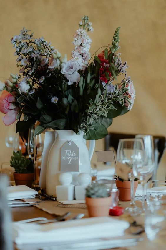 a cute and romantic wedding centerpiece of a white porcelain jug with pastel blooms and greenery and potted succulents around