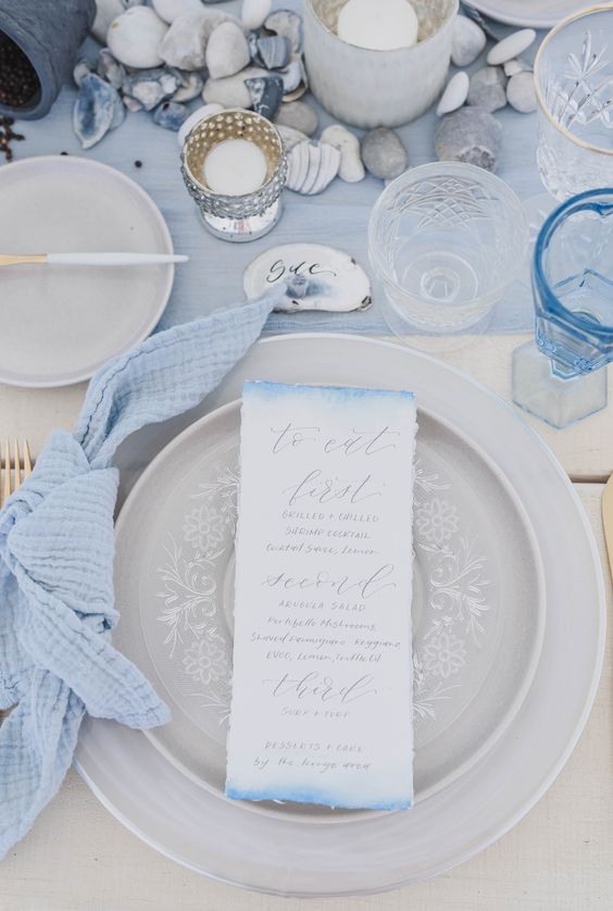 a dreamy ocean-inspired tablescape with blue linens, pebbles, candles, grey lace plates and an ombre menu