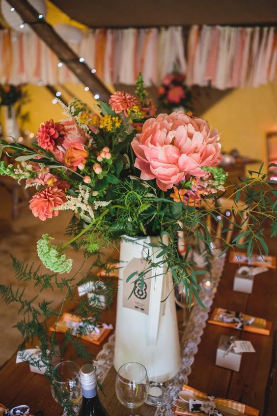 a gorgeous wedding centerpiece of a cream jug with a cardboard tag, pink and orange blooms, greenery and berries for a rustic wedding