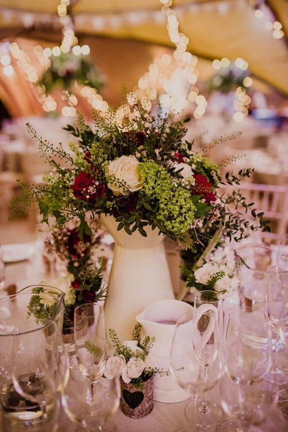 a lush boho wedding centerpiece of a cream jug, bright blooms and white ones, greenery and candles around is amazing for summer or fall