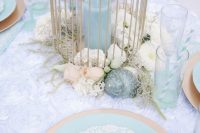 a pretty mermaid bridal shower tablescape with a neutral runner, gold and turquoise plates, a cage lantern with blooms and seashells