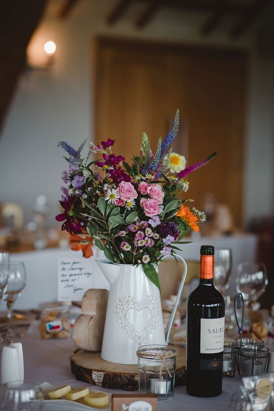 a pretty rustic wedding centerpiece of a wood slice, a metal jug with a heart and bold boho blooms and greenery