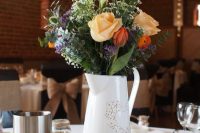 a rustic summer wedding centerpiece of a tree stump, a wood slice with a name, a whit e jug with bold blooms and greenery and a jug