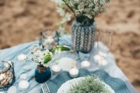 a seaside bridal shower tablescape with blue linens, a blue embroidered placemat, neutral blooms, pebbles and candles
