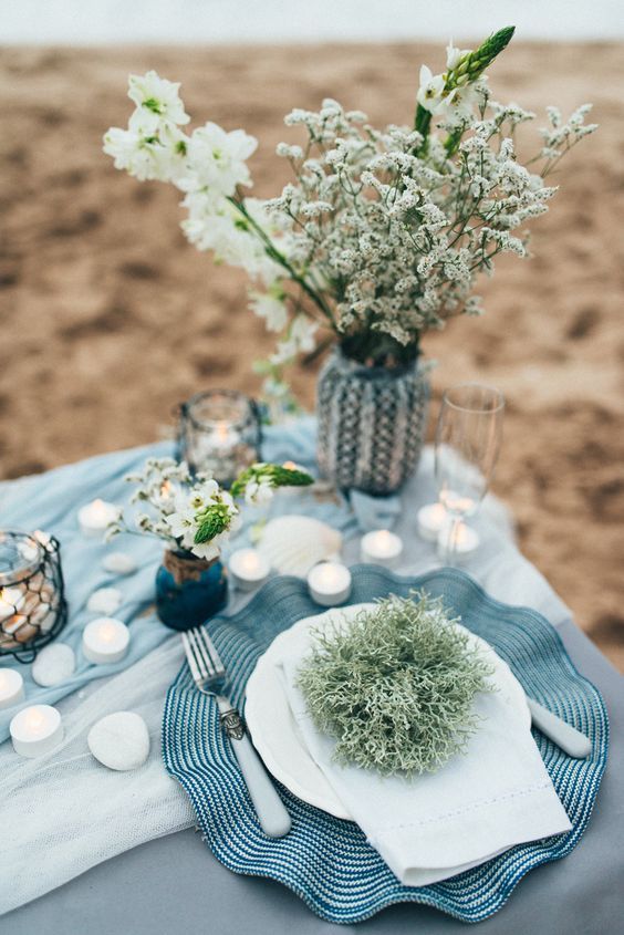 a seaside bridal shower tablescape with blue linens, a blue embroidered placemat, neutral blooms, pebbles and candles