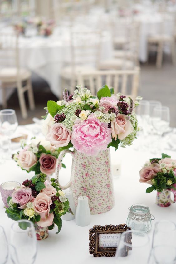 a summer wedding centerpiece of a floral jug with pink and blush blooms plus greenery and matching arrangements around