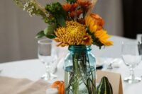 a blue mason jar filled with season flowers of football mums, sunflowers, hardy mums, wheat, open roses, and foliage plus gourds