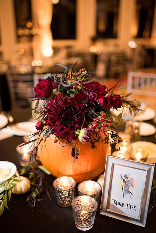 a bold fall wedding centerpiece of an orange pumpkin with burgundy blooms and berries, greenery and candles is wow