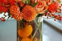 a bold orange fall wedding centerpiece of a clear vase with tiny pumpkins, lush dahlias and roses and more pumpkins and berries