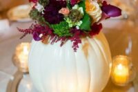 a creative fall wedding centerpiece of a large white pumpkin as a vase for a bold floral arrangement and candles around it