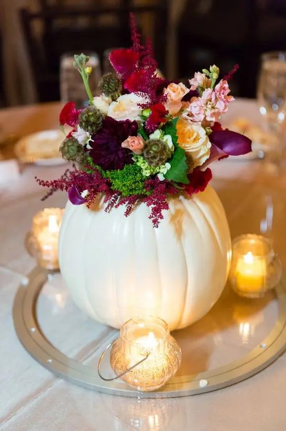 a creative fall wedding centerpiece of a large white pumpkin as a vase for a bold floral arrangement and candles around it