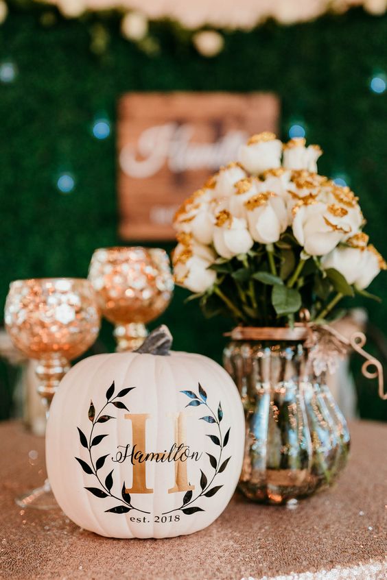a fall wedding centerpiece of white roses with gold glitter in a vase, a white pumpkin with a letter and other decor