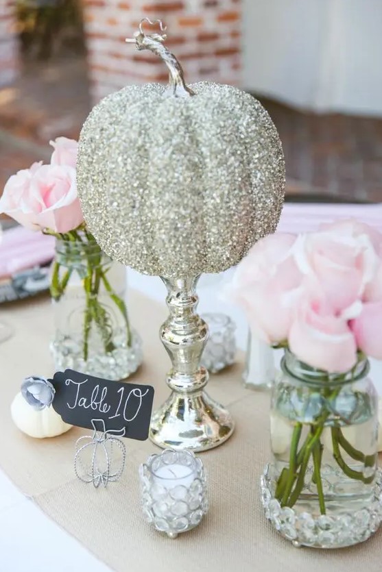 a glam cluster wedding centerpiece of jars with pink roses, a glitter pumpkin on a stand, candles and a chalkboard table number