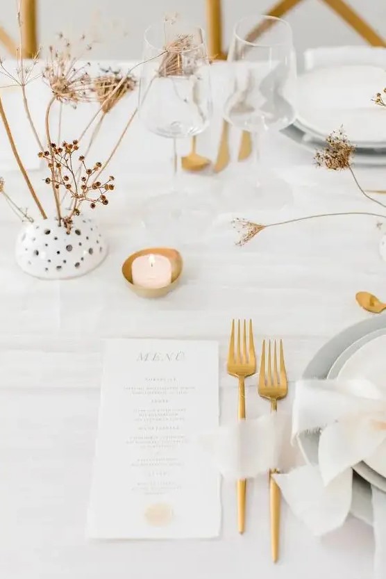 a minimalist neutral wedding table setting with white linens, grey plates and chargers, candles, gold cutlery and dried blooms