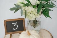 a neutral rustic centerpiece of a wood slice, a white pumpkin, white blooms in a jar and a chalkboard table number