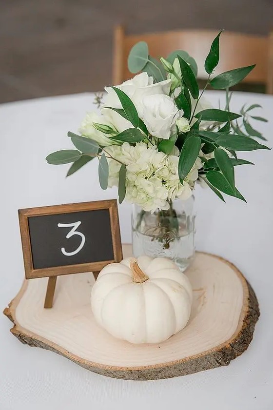a neutral rustic centerpiece of a wood slice, a white pumpkin, white blooms in a jar and a chalkboard table number
