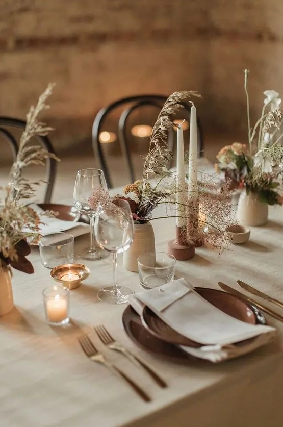 a relaxed boho wedding table setting with neutral linens, terracotta plates, elegant vases with bold blooms and dried grasses and candles