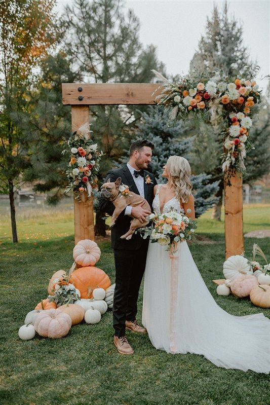 a rustic fall wedding arch decorated with neutral and bright blooms, with stacked pumpkins at the base looks nice