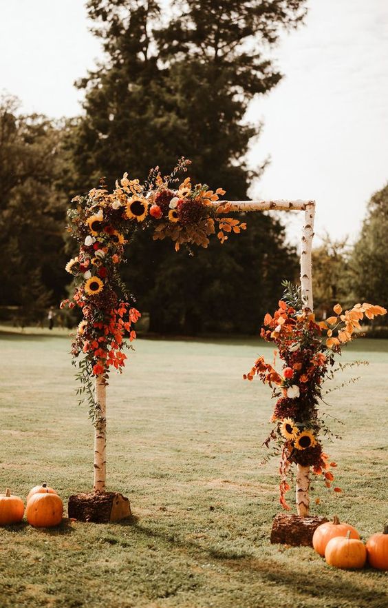 a rustic fall wedding arch with bold blooms, bold leaves and greenery and pumpkins at the base of the arch