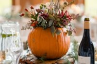 a rustic fall wedding centerpiece of a pumpkin with bold blooms and greenery, a tree slice and fall leaves around is chic