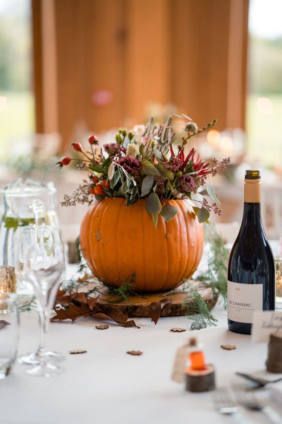a rustic fall wedding centerpiece of a pumpkin with bold blooms and greenery, a tree slice and fall leaves around is chic