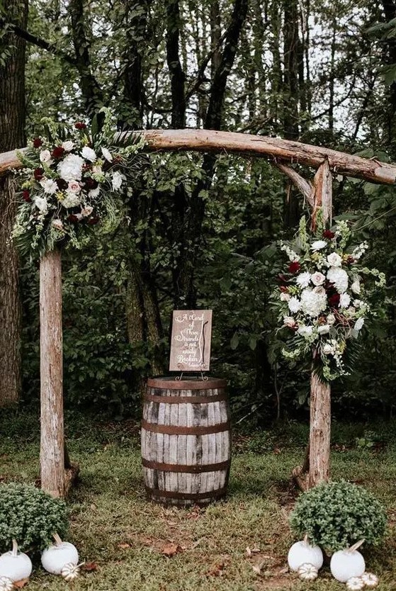 a simple rustic wedding arch with a curved top, of logs, white and burgundy blooms and greenery and white pumpkins on the ground